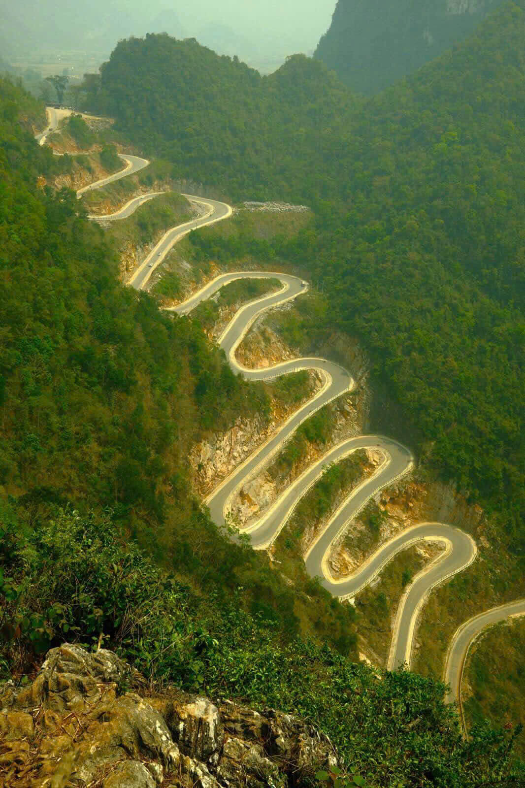 A breathtaking vertical landscape view of the legendary Khau Coc Cha Pass in Cao Bang, Vietnam. The steep, winding mountain road features 14 sharp hairpin turns carved perfectly into the dense, green jungle mountainside.