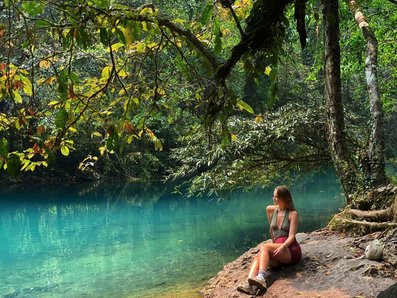 A female traveler sitting peacefully on a rocky bank beside the incredibly clear, turquoise waters of the Lenin River (Suối Lê-nin) at the Pac Bo historical site. She is surrounded by a lush, green jungle canopy with dappled sunlight reflecting off the emerald water, highlighting the serene and untouched beauty of the Cao Bang Loop.