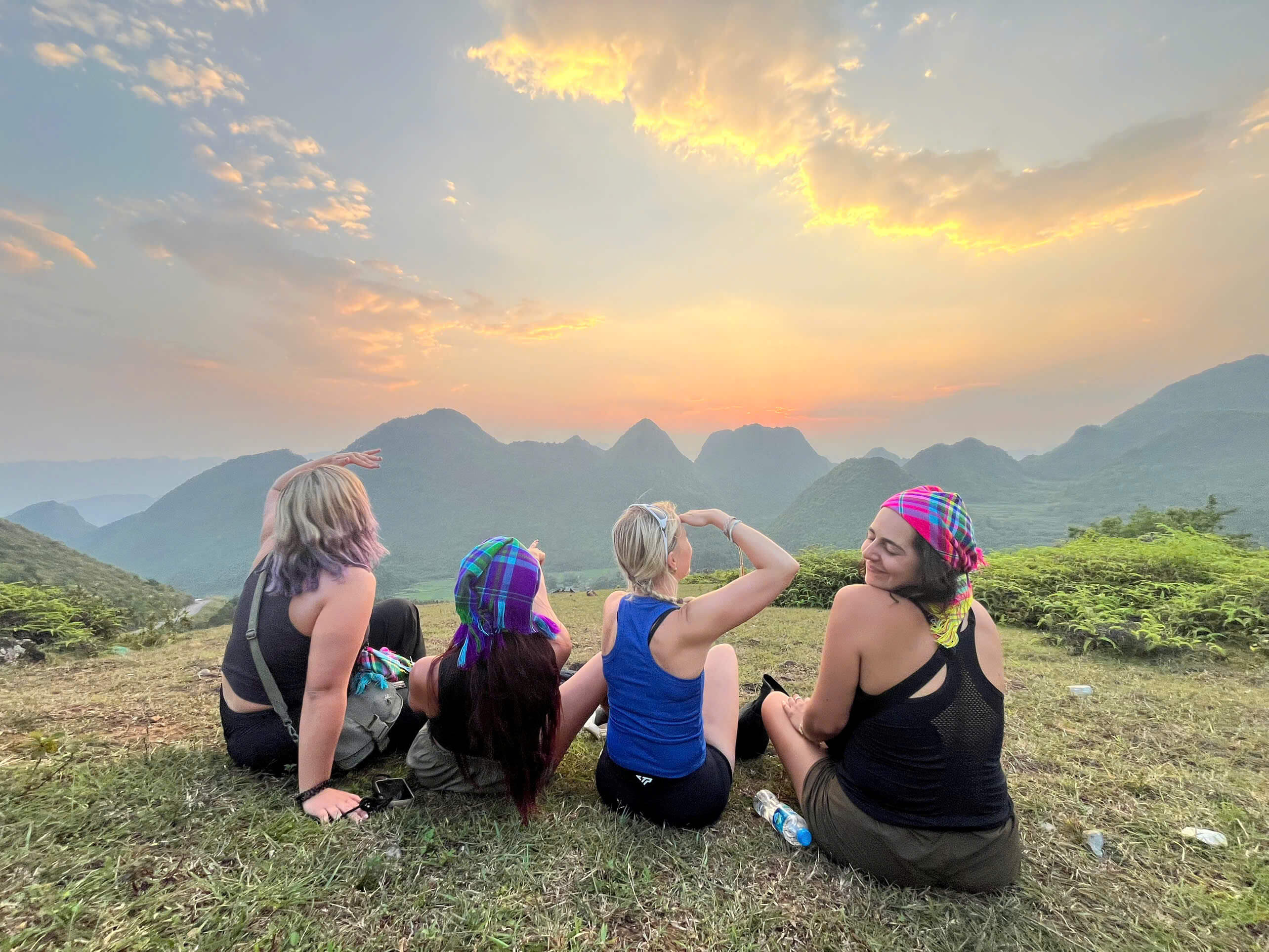 Four female backpackers sit on a grassy hilltop viewpoint, overlooking a dramatic and stunning landscape of interlocking karst mountains and green valleys during a beautiful sunset. Two of the women wear colorful scarves, and the sky is filled with soft orange clouds.