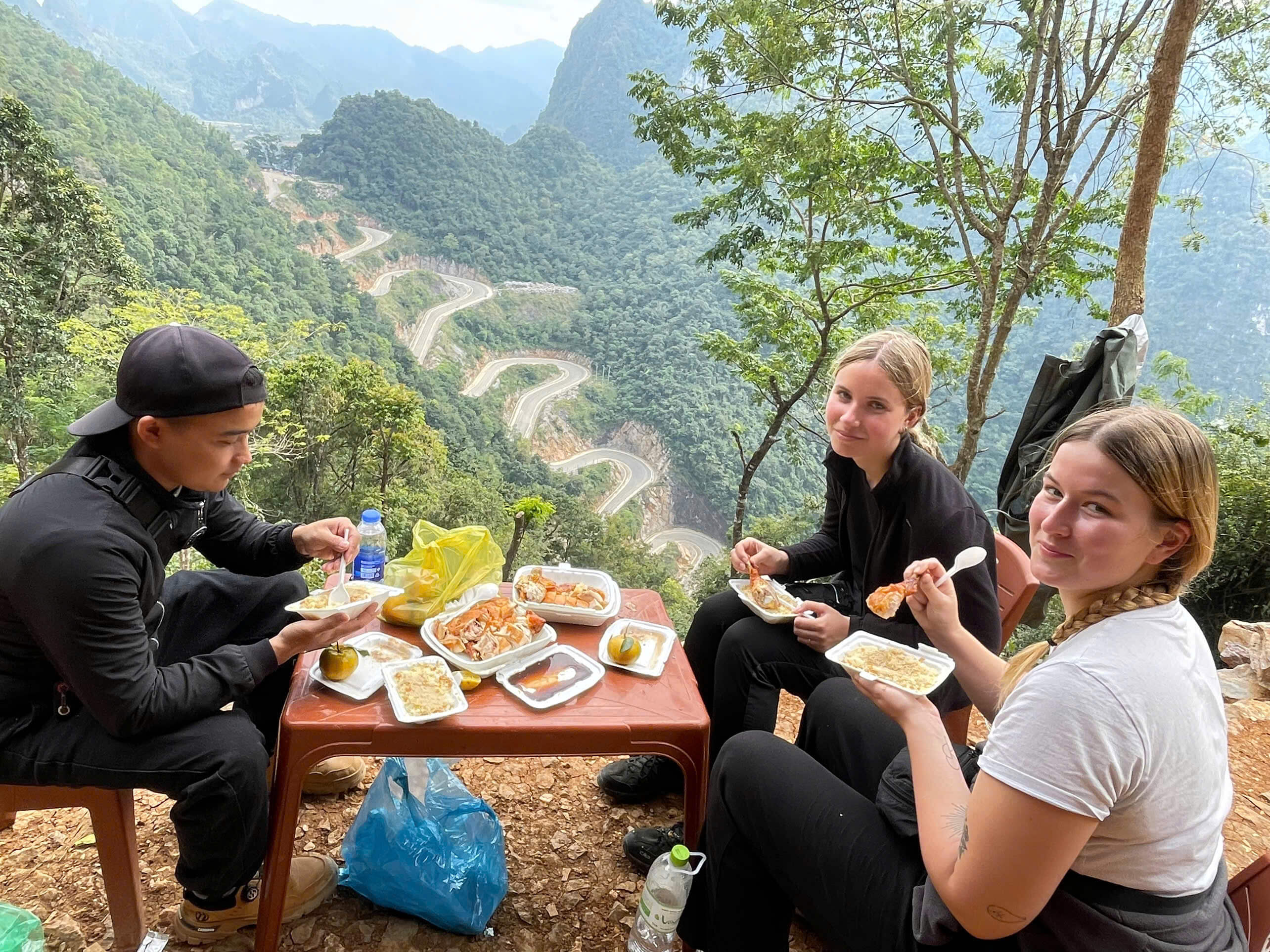 Enjoying a delicious local picnic lunch (featuring roast duck) right at the edge of the mountain. They are resting at a scenic spot that perfectly overlooks the magnificent 14-tier Khau Coc Cha Pass.