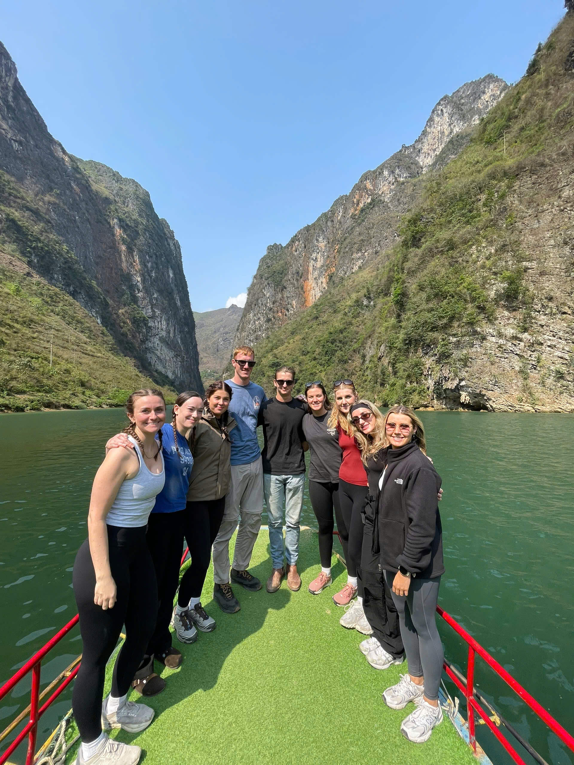 A large group of happy Motorventures travelers posing together on a boat deck, cruising through the majestic Tu San Canyon on the emerald green Nho Que River in Ha Giang.